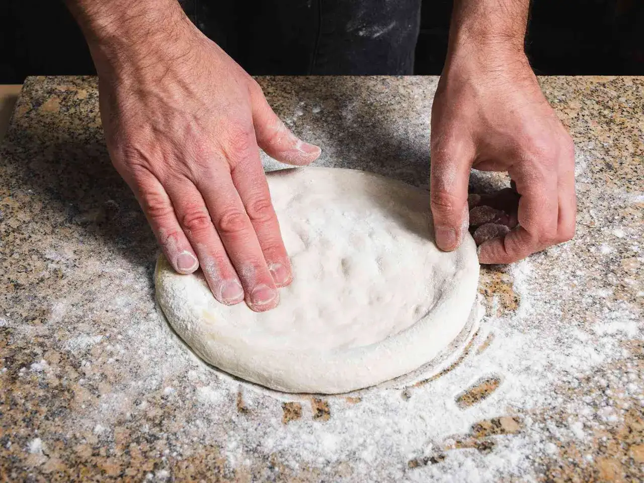 Fredrik formar pizzadeg för en mysig fika. Händerna arbetar varsamt med det mjöliga deget på en stenig bänk.