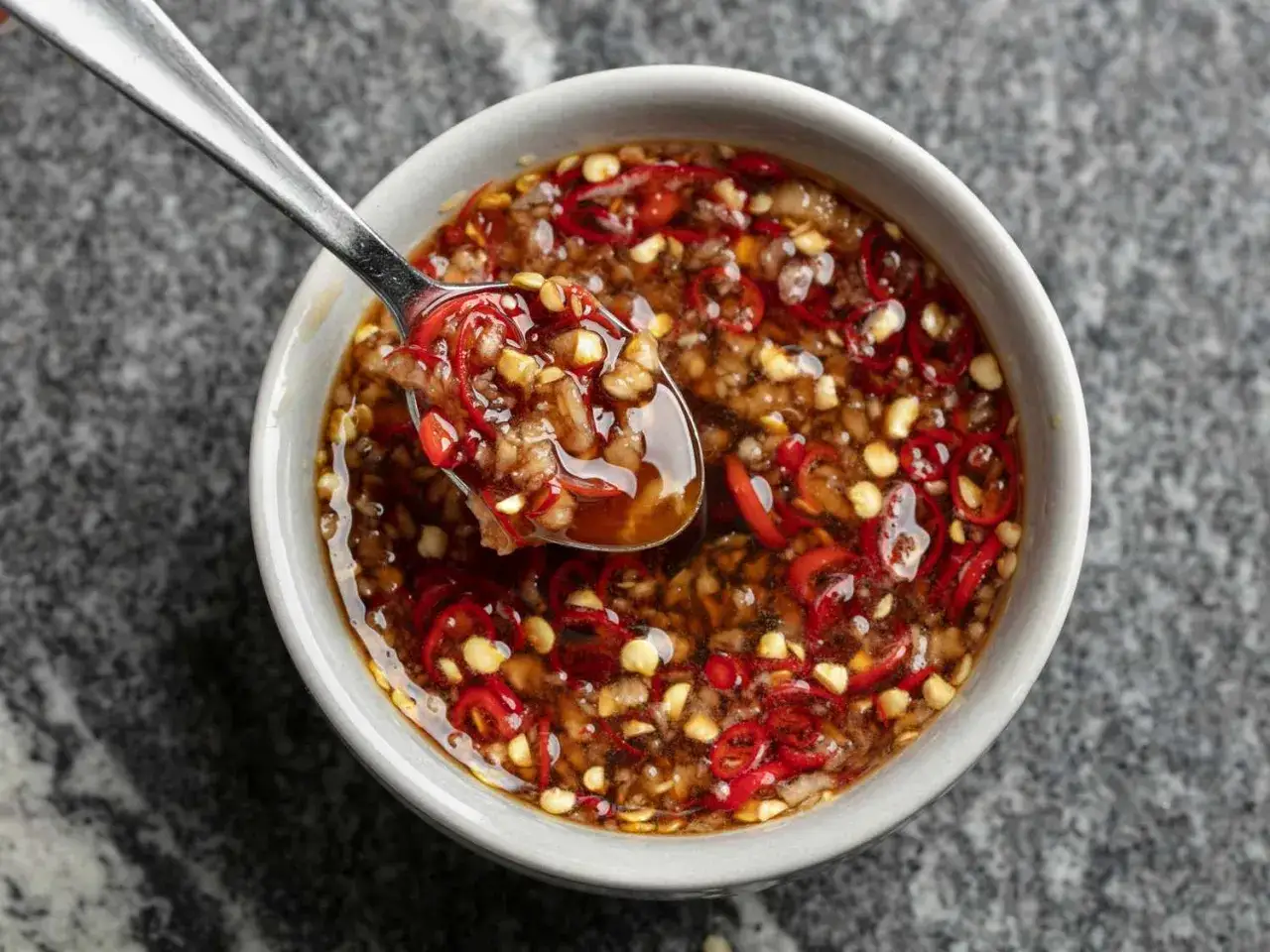 En sked lyfts ur en skål med kryddig sweet chili sås, full av finhackad chili och vitlök.