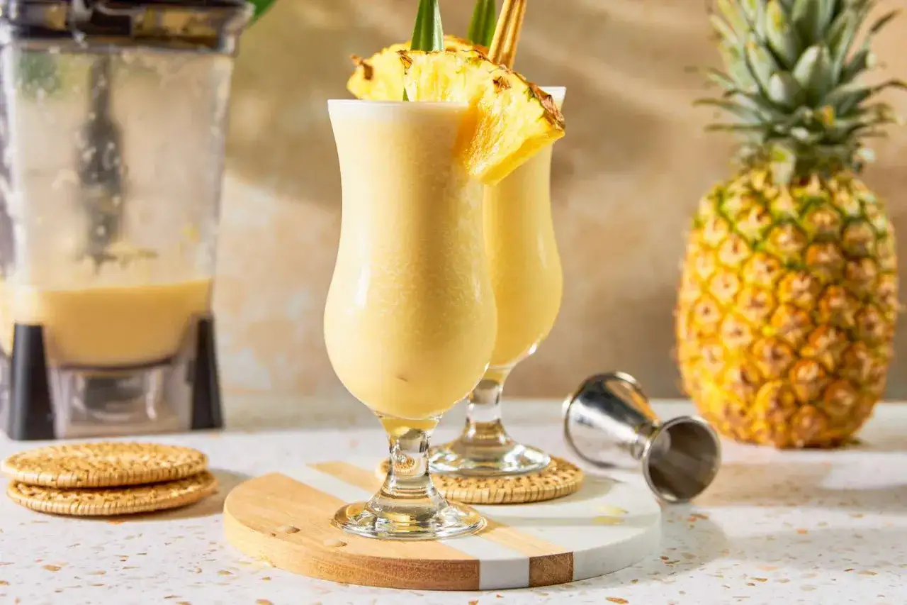 Två glas med krämig Piña Colada, garnerade med ananasskivor. En mixer och en hel ananas syns i bakgrunden. Perfekt för ett pina colada recept.