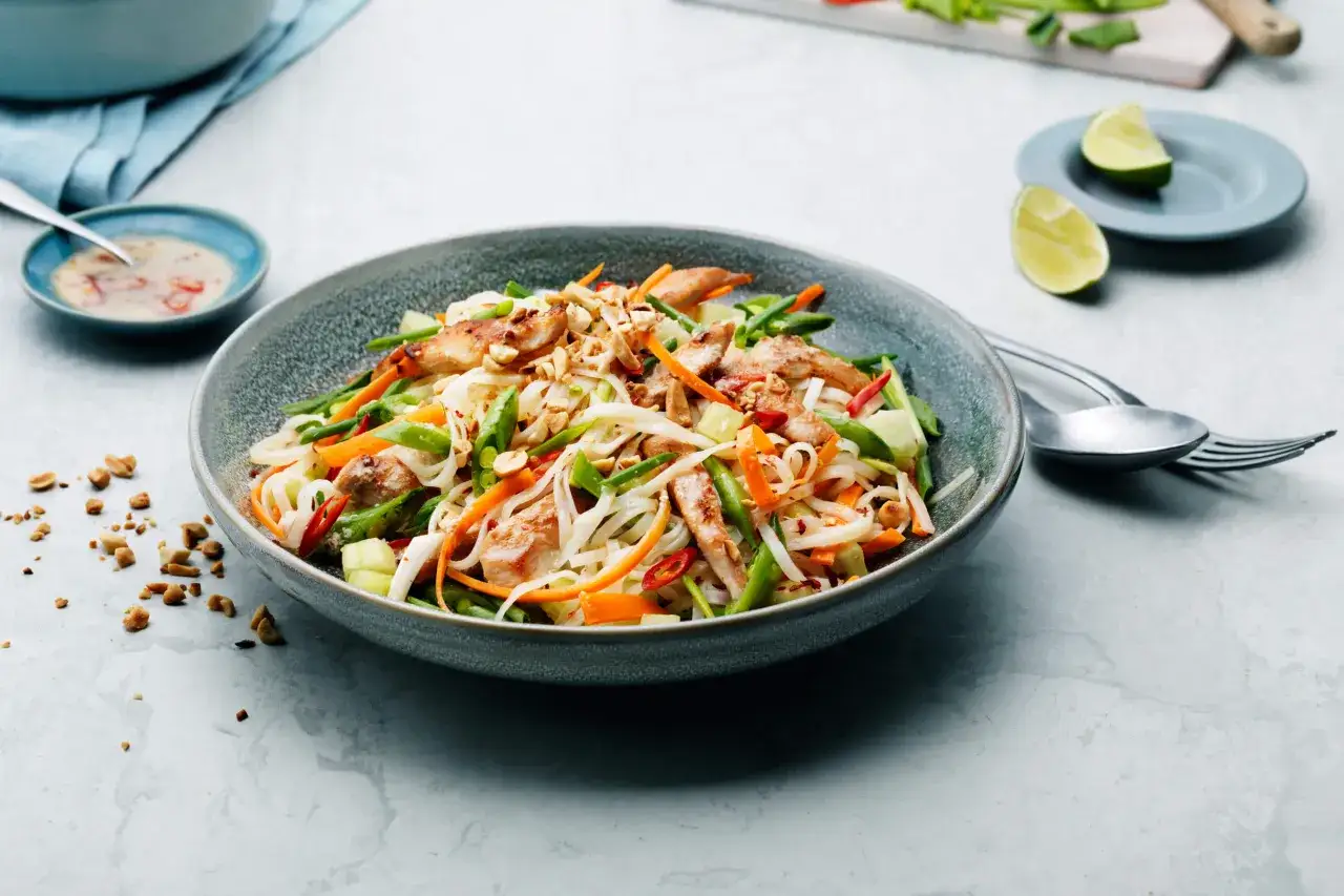 En läcker asiatisk nudelsallad med kyckling, grönsaker och jordnötter. Serveras med en dressing och limeklyftor.