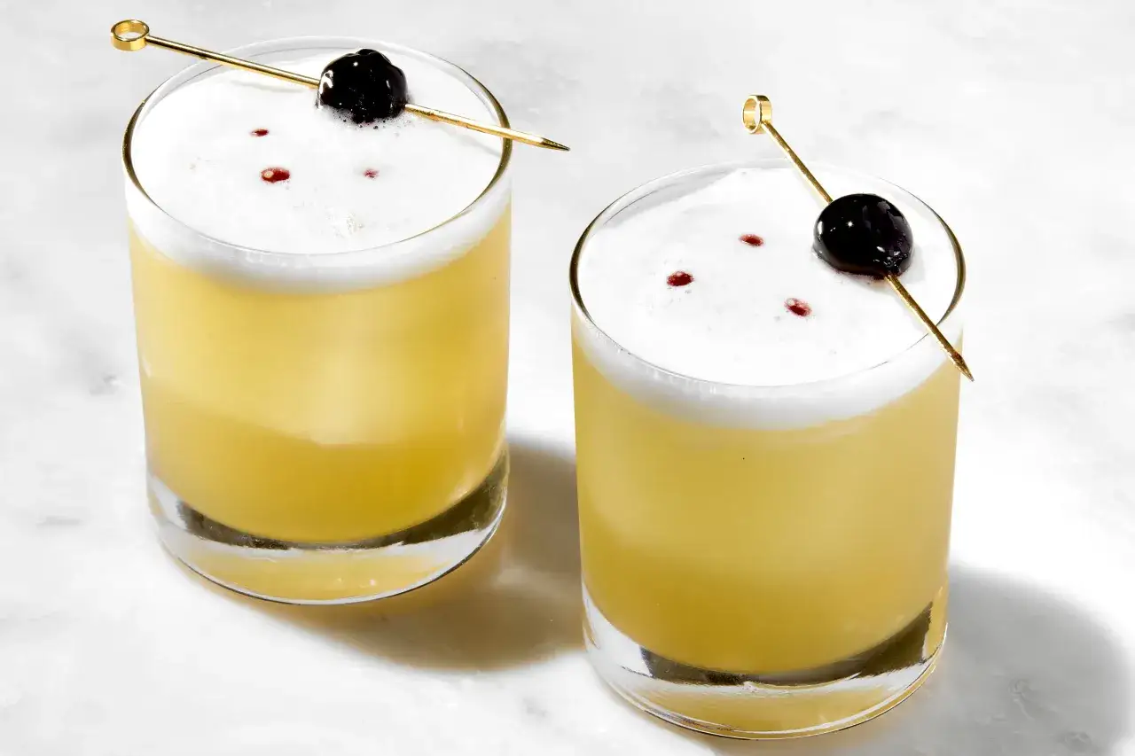 Två glas med en gyllene whiskey sour, toppad med vitt skum och en körsbärsdekoration.